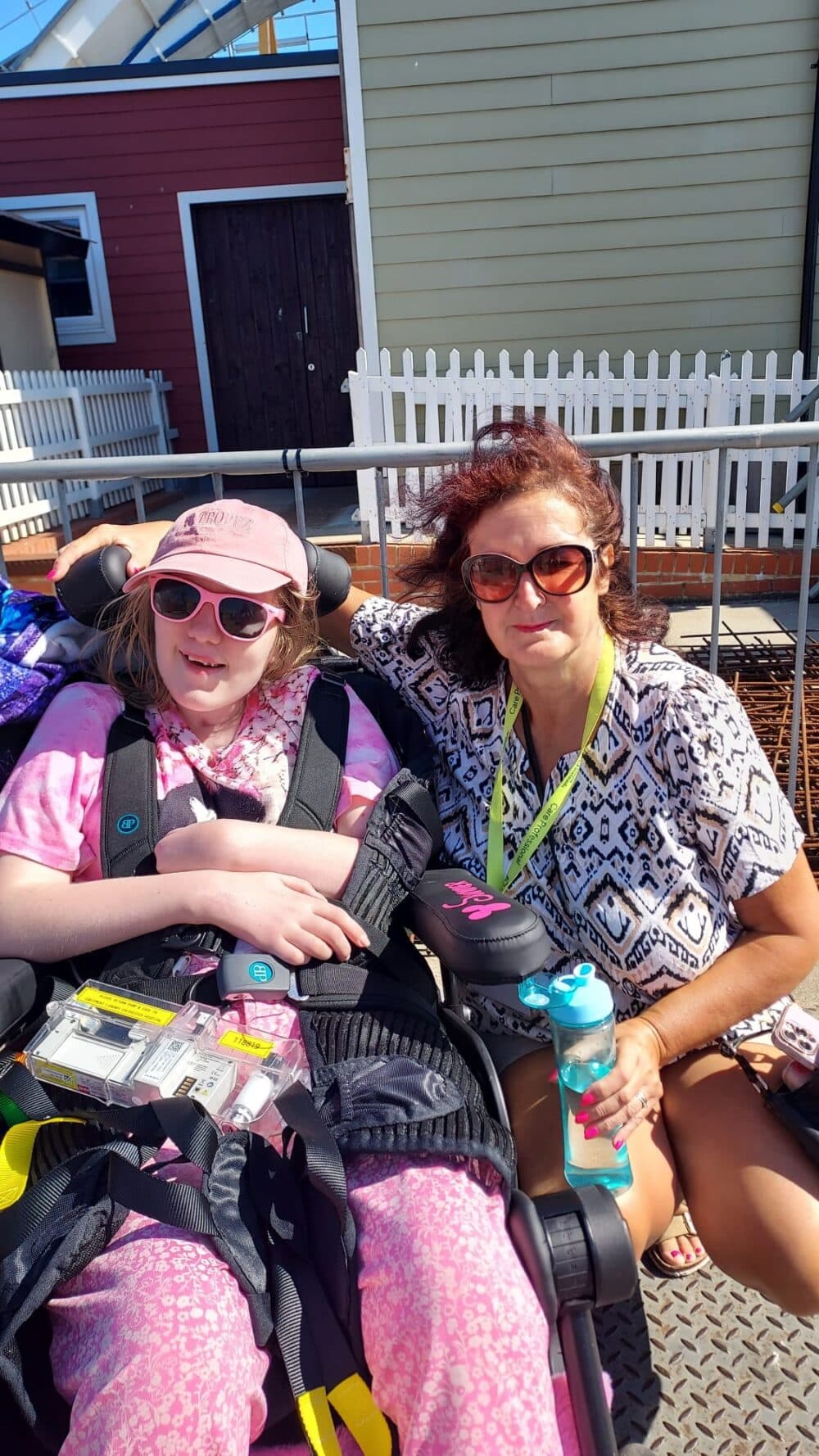 A woman and a girl in a wheelchair sit outdoors, both smiling and wearing sunglasses on a sunny day. - Home Instead