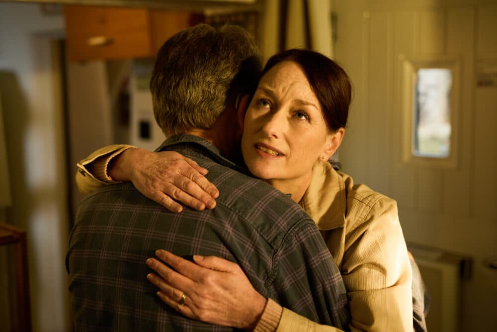 A woman hugs a man and looks upwards with a thoughtful expression in a warmly lit room. - Home Instead