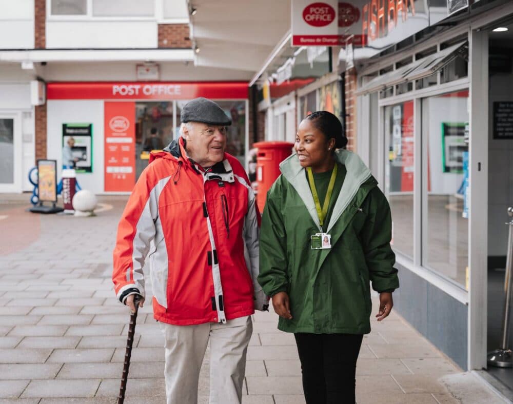 An elderly man with a walking stick walks beside a smiling woman outside a post office on a city street. - Home Instead