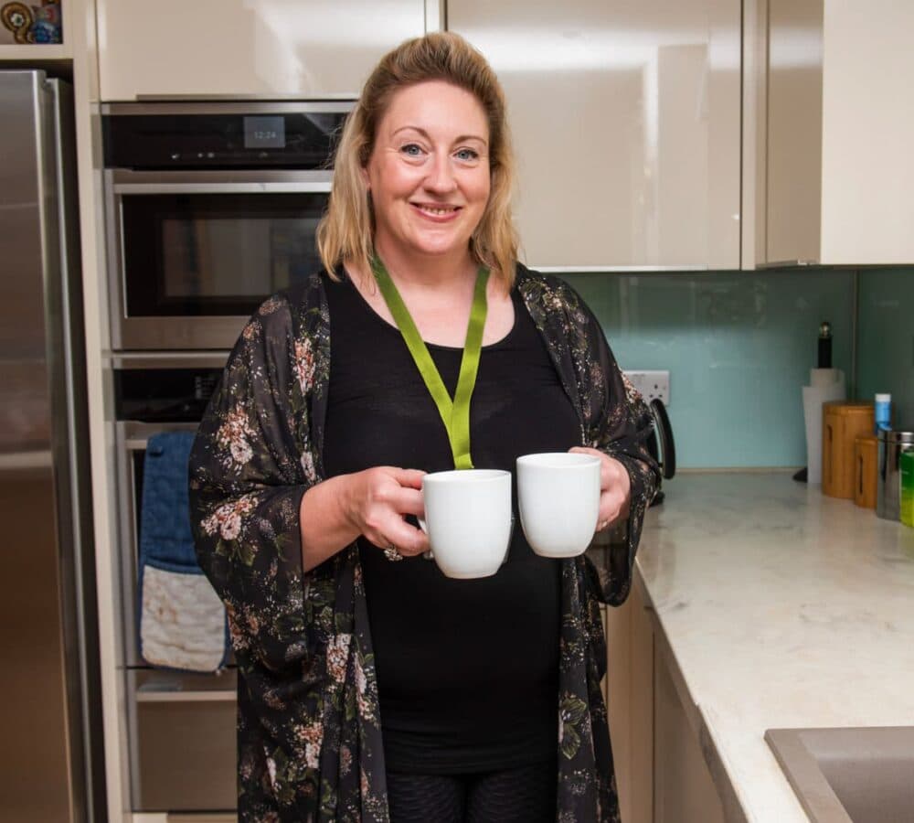 A woman with short blonde hair holding two mugs while smiling and standing