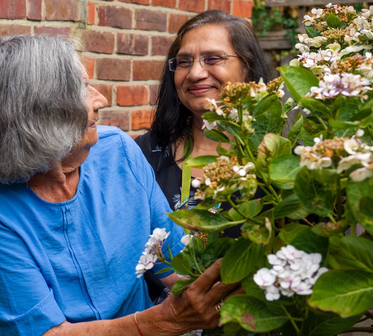 Two women smiling and talking beside a flowering shrub in front of a brick wall. - Home Instead