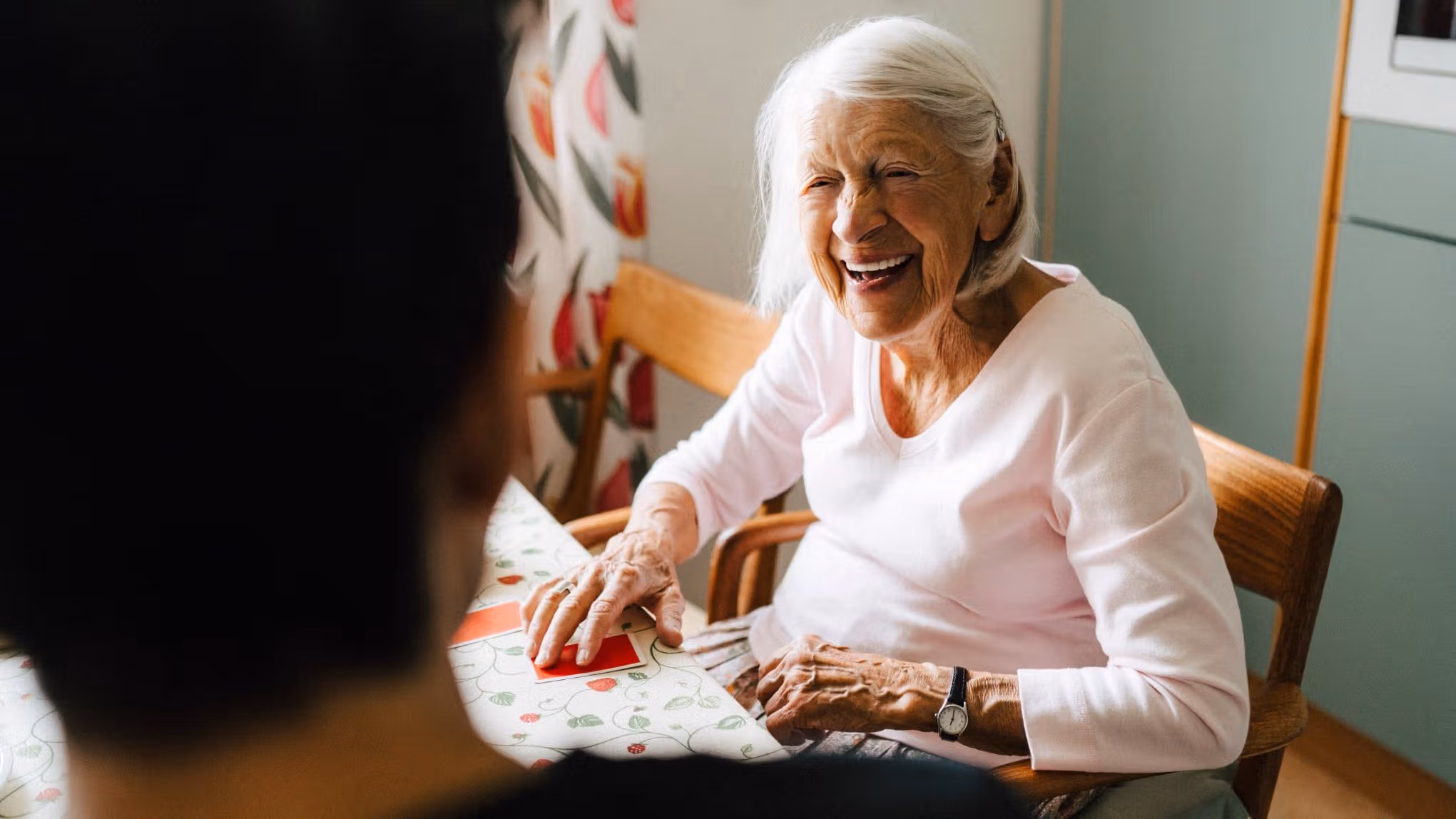 Smiling elderly woman sitting at a table, talking with another person in a cosy room. - Home Instead