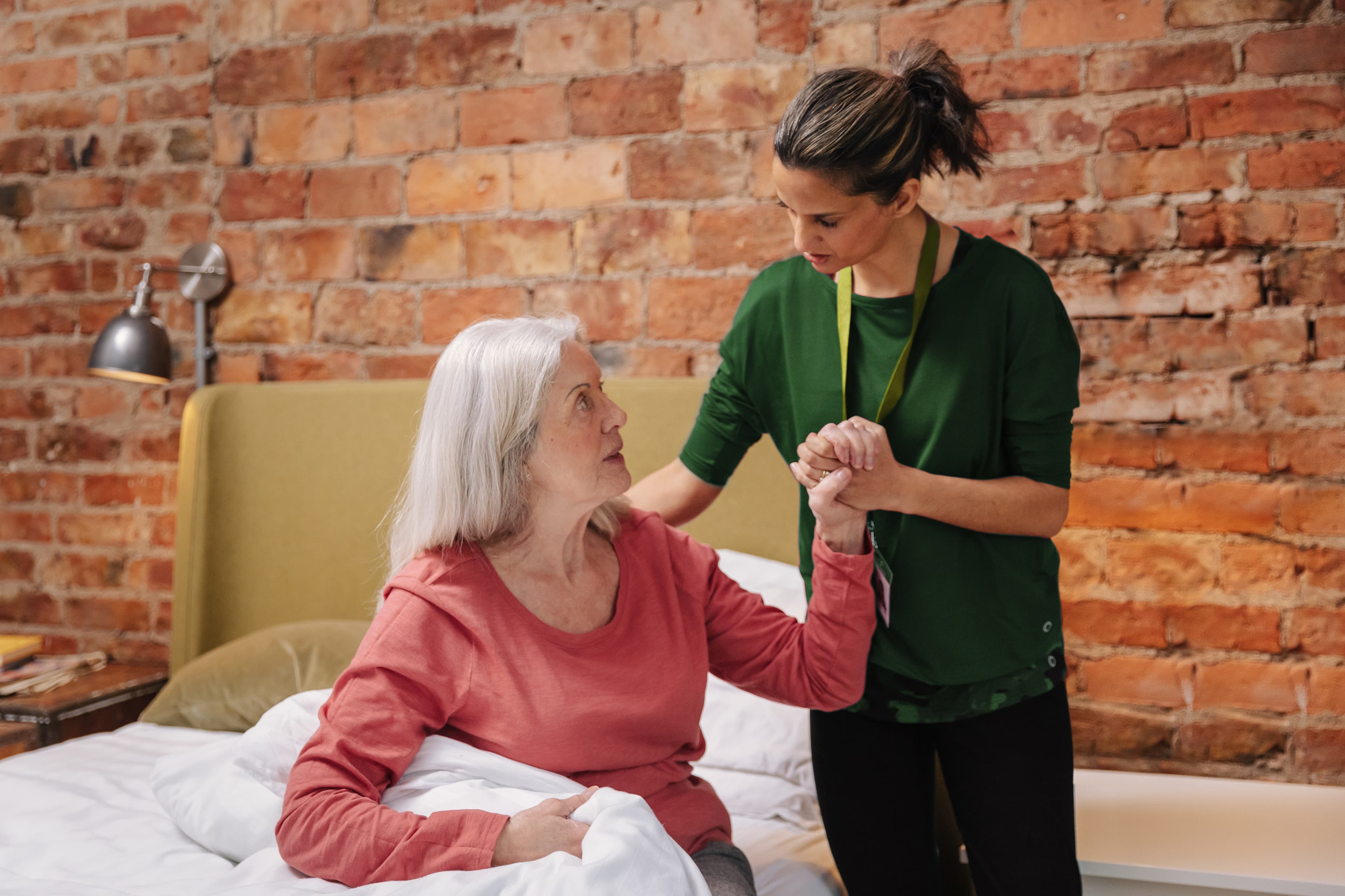 Carer helps an elderly woman sit up in bed against a brick wall background in a cosy room. - Home Instead