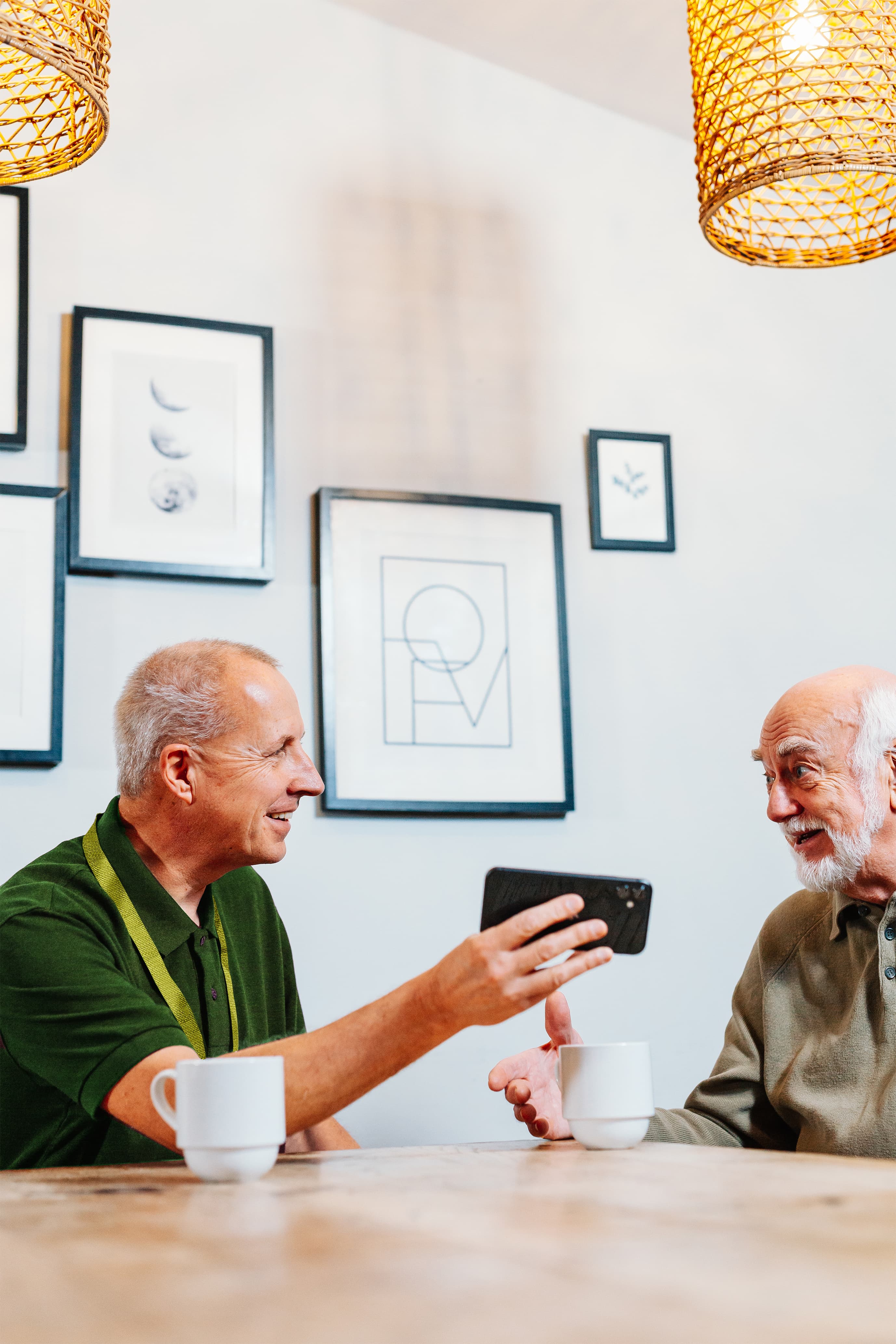 Two older men sit at a table with coffee, smiling and looking at a mobile phone in a cosy café setting. - Home Instead