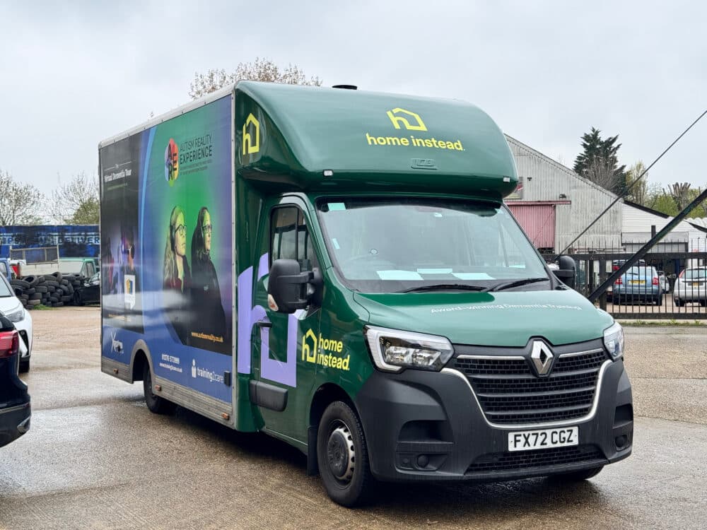 A green Home Instead van with branding and images, parked in an outdoor car park near industrial buildings. - Home Instead