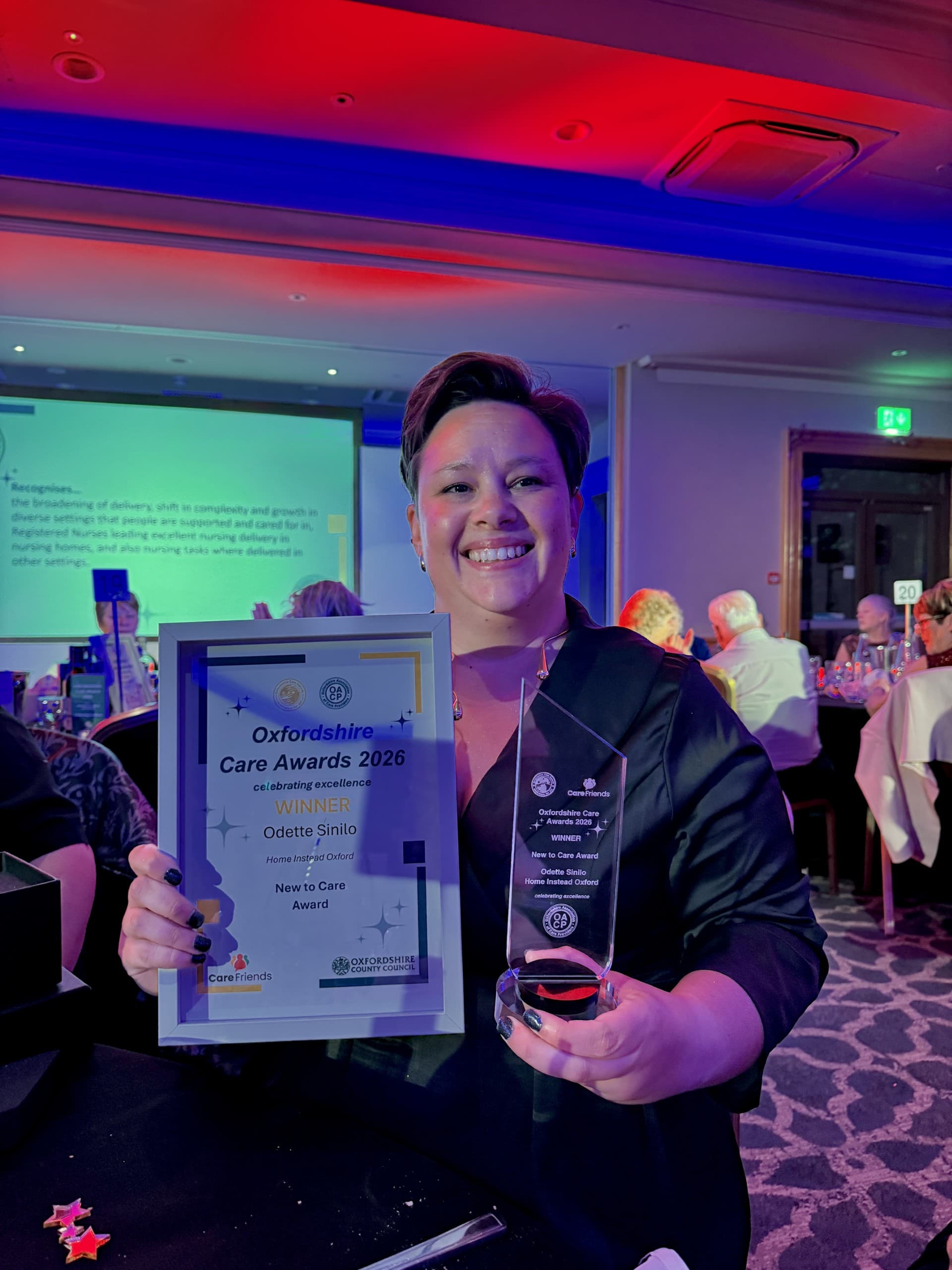 Smiling person holding a framed award and trophy at a formal event with people seated at tables. - Home Instead