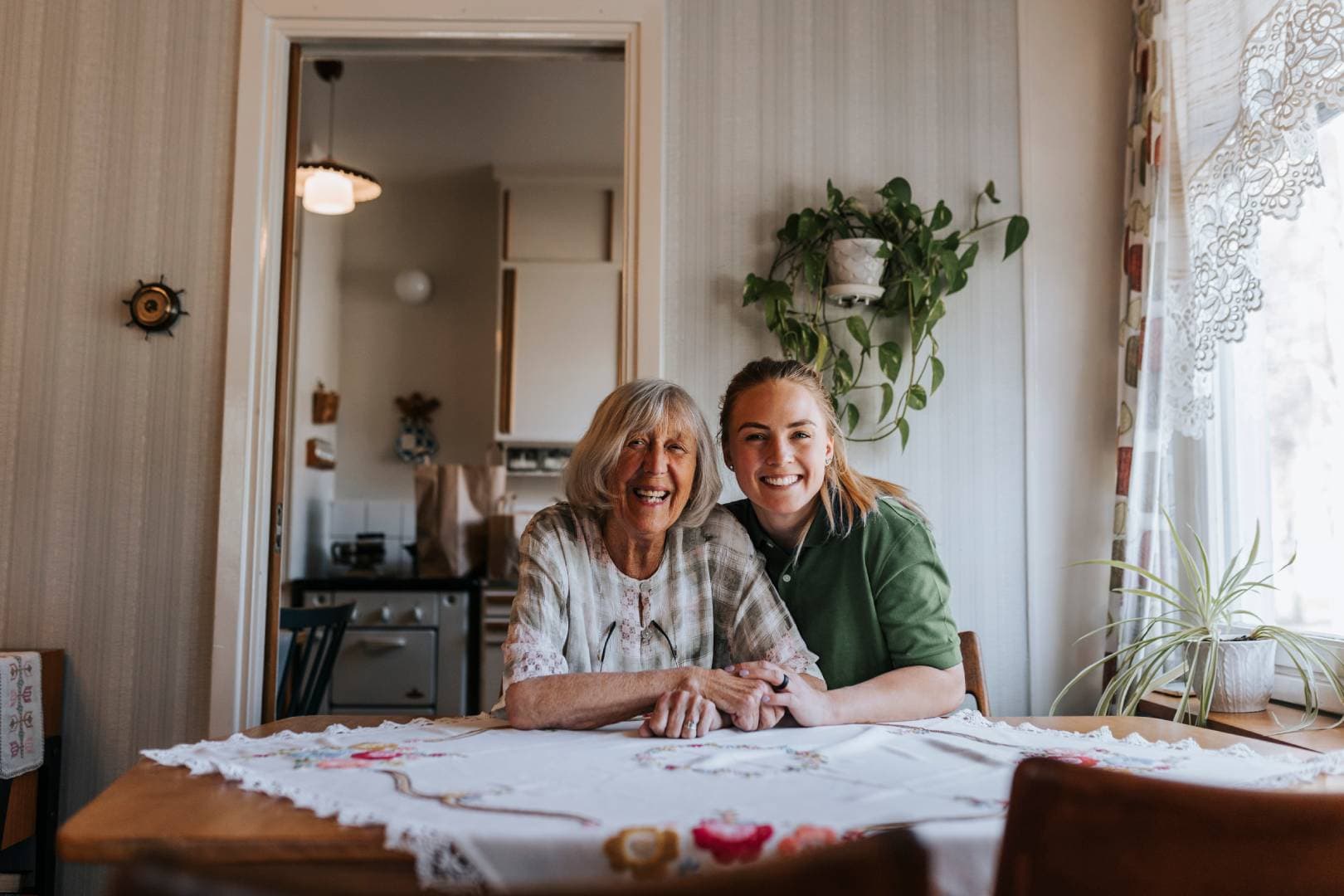 Two women smiling and sitting together at a kitchen table, holding hands in a cosy, sunlit room. - Home Instead