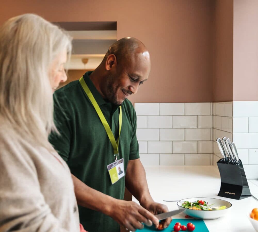 A male carer wearing green happy and smiling while chopping vegetables with an older female adult