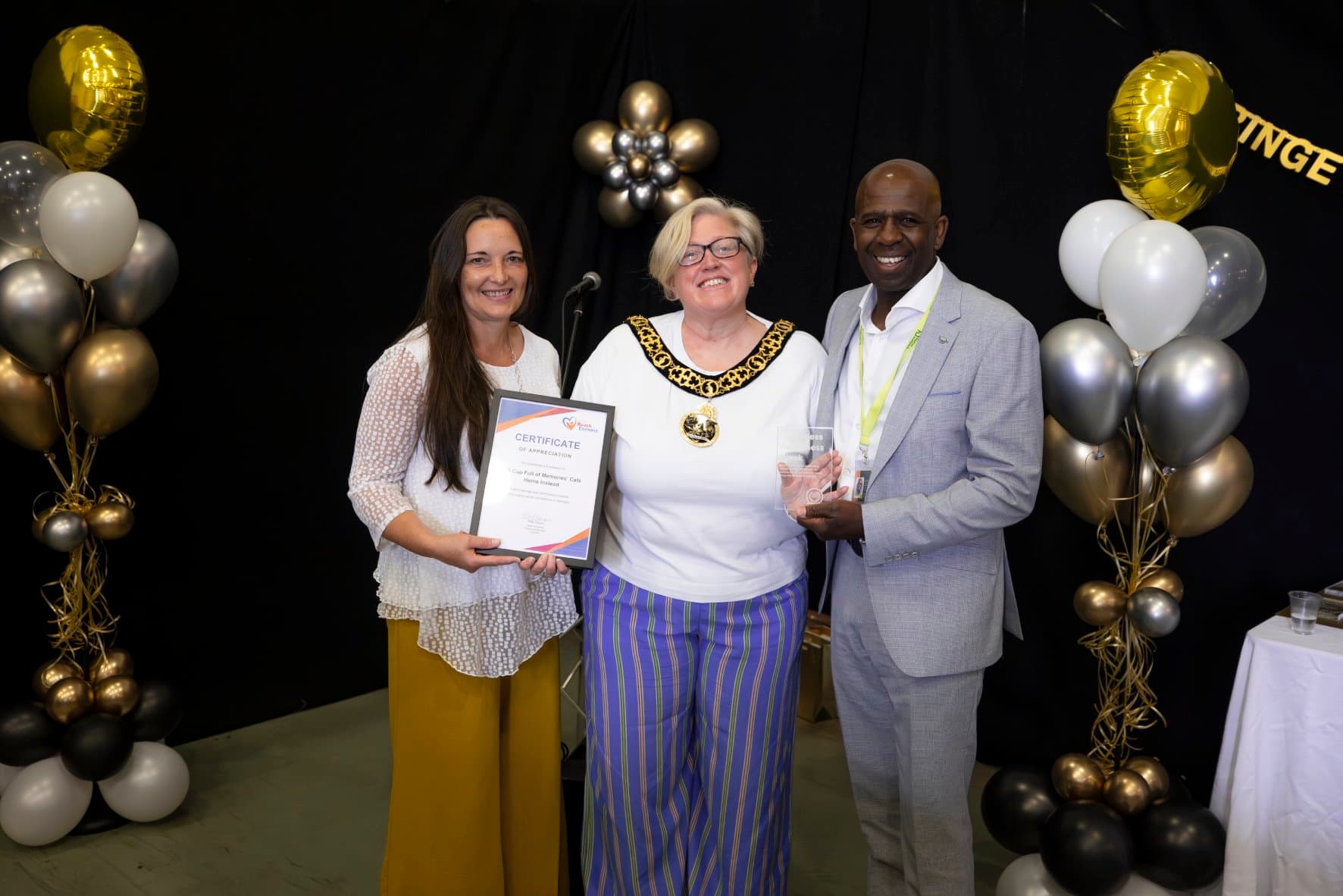 Three people smiling, holding a certificate and trophy, stand between balloon displays at an awards ceremony. - Home Instead