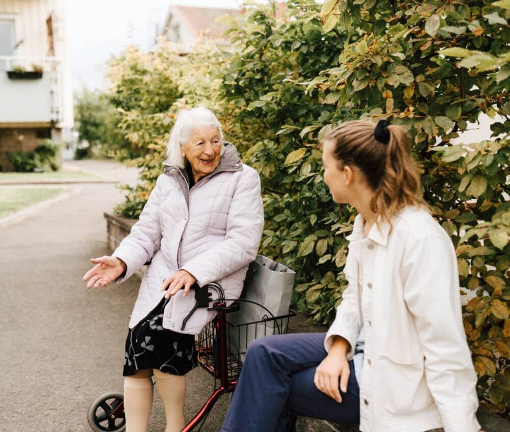An elderly woman with a walking frame smiles and talks with a younger woman on a pavement near greenery. - Home Instead